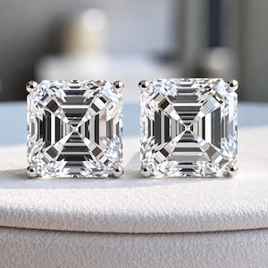 May include: A pair of square-cut stud earrings featuring clear, faceted stones set in a four-prong silver-toned metal setting. The stones exhibit a distinct step-cut pattern with sharp geometric lines and high brilliance, reflecting light clearly.