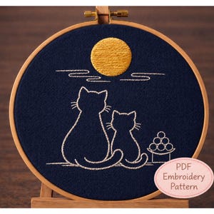 May include: Embroidery hoop art featuring two cat silhouettes gazing at a golden moon against a navy blue background. The design includes white stitched details of clouds and a small offering. The text "PDF Embroidery Pattern" is also included.