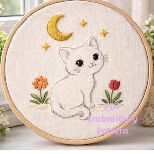 以下が含まれることがあります： 木製のフレームに刺繍された猫のデザイン。白い猫が三日月と星の下に座っています。猫の両側に花があります。「PDF Embroidery Pattern」の文字が下にあります。