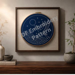 May include: A framed embroidery hoop featuring a navy blue design with a crane, moon, and stars. The design is stitched with white thread, depicting waves at the bottom. The text "DE Embroidery Pattern" is visible. The frame is dark brown, and the background is a neutral linen.