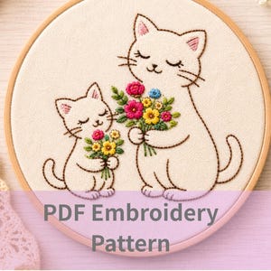 以下が含まれることがあります： カラフルな花束を持った2匹の白い猫の手刺繍デザインの刺繍枠。 猫は目を閉じています。 画像の下部には「PDF Embroidery Pattern」というテキストがあります。