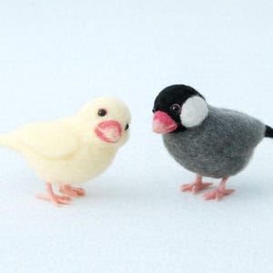 以下が含まれることがあります： 2つのフェルト鳥の置物。1つはピンクのくちばしと足を持つ淡い黄色です。もう1つは、黒い頭、白い頬のパッチ、ピンクのくちばしを持つグレーです。どちらの鳥も小さな黒い目を持ち、立っています。