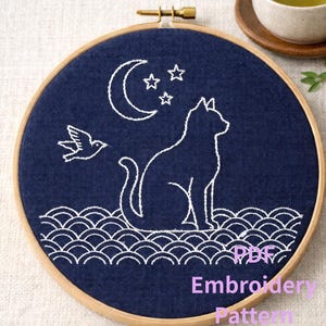 Può includere: Telaio da ricamo con tessuto blu navy con un ricamo bianco di un gatto, luna, stelle e uccello. Il design include motivi a onde nella parte inferiore. Il testo "PDF Embroidery Pattern" è visibile.