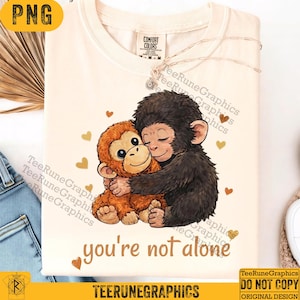 Puede incluir: Camiseta color crema con un gráfico de dos monos de dibujos animados abrazándose, rodeados de corazones dorados. El texto "you're not alone" está impreso debajo. Un collar plateado está sobre la camiseta.