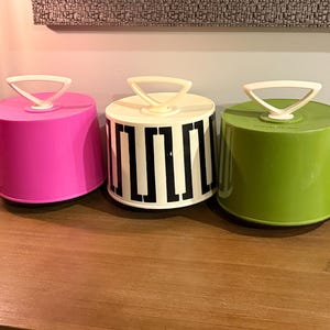 Puede incluir: Tres portatartas redondos con asas triangulares blancas. El de la izquierda es de color rosa intenso, el central es blanco con un patrón geométrico griego negro y el de la derecha es de color verde lima. Cada uno tiene una tapa plana.