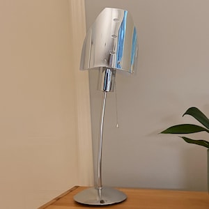 May include: A contemporary desk lamp with a chrome finish, featuring a curved stem and a circular base. The lamp shade is silver and reflective, with three small circular openings. A pull chain is visible. The lamp is on a wooden surface.