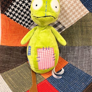 Puede incluir: Un peluche verde lima con forma de lagarto. Tiene ojos blancos grandes, una cresta azul y parches de cuadros vichy rosas y cuadros azules. El juguete tiene un apéndice marrón en forma de rama.
