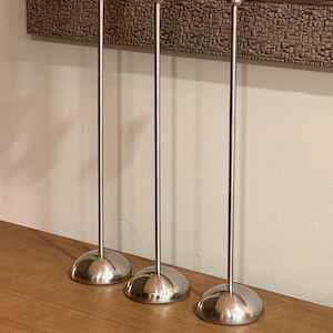 Può includere: Tre candelabri argentati dal design minimalista. Ognuno ha una base rotonda, un gambo sottile e una coppa per una candela bianca. I candelabri sono disposti su una superficie di legno. Gli steli sono alti circa 25 cm.