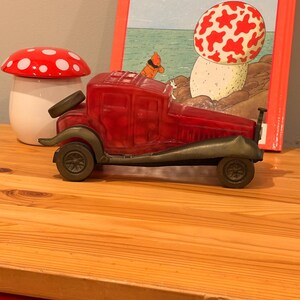 Peut inclure: Un camion jouet rouge avec des accents couleur bronze est posé sur une surface en bois, à côté d'un pot en forme de champignon avec un couvercle à pois rouges et blancs. Un livre intitulé "L'Étoile Mystérieuse" est en arrière-plan.