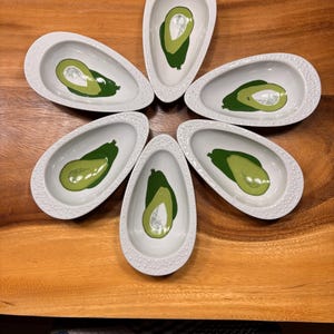 Peut inclure: Six petits plats en céramique blancs en forme d'avocat, disposés en cercle. Chaque plat présente un motif d'avocat vert avec un noyau argenté. Les plats ont un bord texturé et pointillé et sont posés sur une surface en bois.