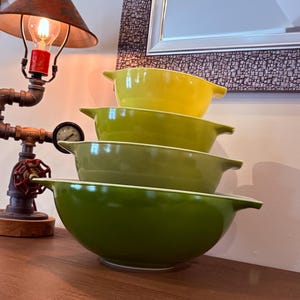 May include: A stack of four vintage Pyrex mixing bowls in shades of green and yellow. The bowls have a classic design with a rounded shape and small handles. The bowls are nested on a wooden surface.
