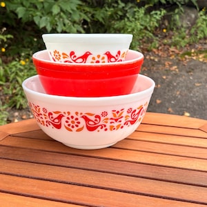 Peut inclure: Une pile de trois bols gigognes. Le bol supérieur est blanc avec un motif d'oiseaux et de fleurs rouges et orange. Le bol du milieu est rouge uni. Le bol inférieur est blanc avec un motif d'oiseaux et de fleurs rouges et orange assorti.