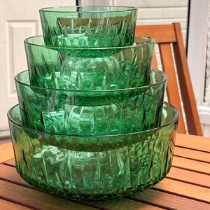 Peut inclure: Ensemble de cinq bols en verre vert de tailles variées, empilés. Les bols présentent un motif texturé en verre taillé. Ils sont disposés sur une table en bois, avec une chaise en bois et une porte blanche en arrière-plan.