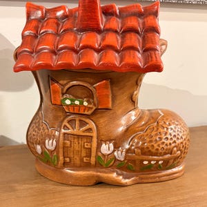 Peut inclure: Une maison en céramique en forme de botte avec un toit et une cheminée rouges. La botte marron présente une porte, une fenêtre avec des volets et des motifs floraux. C'est un objet décoratif.