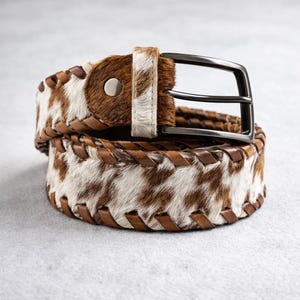 May include: A brown and white cowhide belt with a black metal buckle. The belt features a braided leather edge and a silver-tone button. The cowhide pattern is a mix of brown and white.
