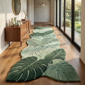 May include: A long, runner-style rug featuring a die-cut shape that mimics a series of overlapping tropical monstera and palm leaves. The rug displays a gradient of green shades, ranging from light sage to deep forest green, with textured leaf veins.