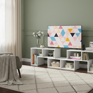 May include: A modern living room setup featuring a white geometric TV stand with open shelving, a gray armchair, and a patterned rug. A large TV displays a colorful geometric design. The room has a neutral color palette.