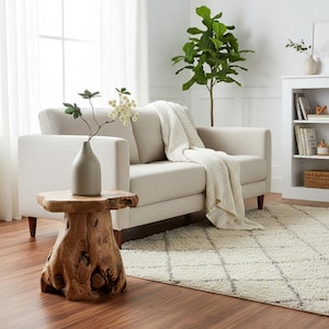 May include: A living room scene featuring a light beige sofa with a cream-colored throw blanket. A natural wood side table supports a gray vase with a floral arrangement. A white rug and a bookshelf complete the decor.