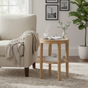 May include: A light wood round side table with a lower shelf. A white mug with steam, an open book, and a small vase with greenery sit on top. A beige blanket drapes over a beige armchair.