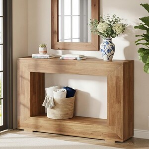 May include: A light brown wooden console table with a rectangular frame. A woven basket with rolled towels sits on the lower shelf. A vase with flowers and a mirror are on the top. A small potted plant and books are also on the top.