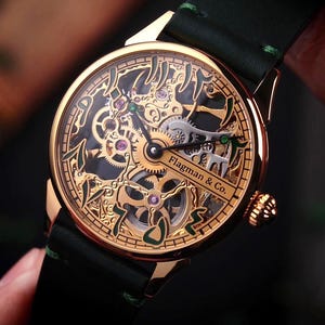 Op de afbeelding: Een goudkleurig skeleton horloge met een zwarte leren band. De wijzerplaat van het horloge toont ingewikkelde tandwielen, paarse juwelen en de tekst "Flagman & Co.". Het horloge heeft een donkergroen accent op de band.
