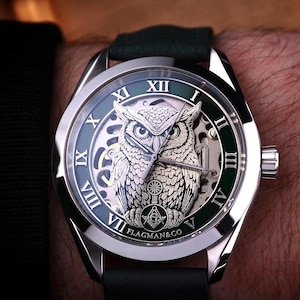 May include: A silver wristwatch with a green and silver face featuring an owl design. The watch has Roman numerals and a black leather band. The brand name "FLAGMAN&CO" is visible at the bottom of the watch face.