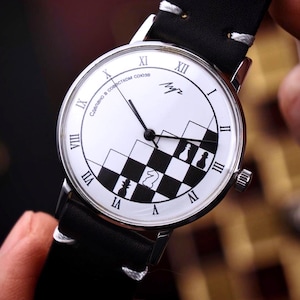 May include: A wristwatch with a white face featuring black Roman numerals and a black and white chess-themed design. The watch has a black leather band and silver casing. The watch face includes the text "Сделано в совести СССР".