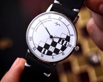 May include: A wristwatch with a white face featuring black Roman numerals and a black and white chess-themed design. The watch has a black leather band and silver casing. The watch face includes the text "Сделано в совести СССР".