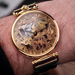 Op de afbeelding: Een goudkleurig polshorloge met een gedetailleerd open-face ontwerp. Het horloge heeft een zwarte leren band en een decoratieve wijzerplaat met ingewikkelde tandwielen en een figuur. Het horloge zit om een pols.