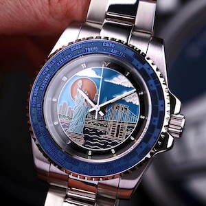 Op de afbeelding: Roestvrijstalen polshorloge met een blauwe lunette met stadsnamen. De wijzerplaat toont een kleurrijke illustratie van het Vrijheidsbeeld, een brug en een zonsopgang. Het horloge heeft een zilveren band.
