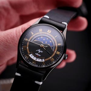 May include: A black wristwatch with a leather strap. The watch face displays gold Roman numerals, a moon phase design, and the Cyrillic text "Луч". The watch has a date display and is held by a person.