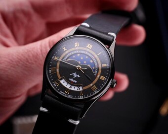 Könnte beinhalten: Eine schwarze Armbanduhr mit Lederarmband. Das Zifferblatt der Uhr zeigt goldene römische Ziffern, ein Mondphasen-Design und den kyrillischen Text "Луч". Die Uhr hat eine Datumsanzeige und wird von einer Person gehalten.