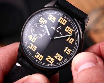 Könnte beinhalten: Eine schwarze Armbanduhr mit einem schwarzen Lederarmband. Das Zifferblatt der Uhr ist schwarz mit goldenen Ziffern und Zeigern. Die Uhr hat ein kleines Hilfszifferblatt und das Wort "Molnija" ist auf dem Zifferblatt aufgedruckt.