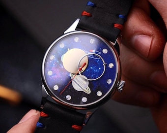 May include: A wristwatch with a celestial design. The watch face features a galaxy background with a planet and a moon. The watch has a black leather strap with red and blue stitching. The watch case is silver.