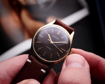 Könnte beinhalten: Eine goldfarbene Armbanduhr mit einem dunkelbraunen Zifferblatt und einem braunen Lederarmband. Die Uhr hat goldene Zeiger und Markierungen, und das Armband ist mit weißem Faden genäht. Die Uhr wird von einer Person gehalten.