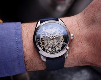 May include: A silver wristwatch with a dark blue leather band. The watch face features an owl design with Roman numerals and the text "FLAGMAN&CO". The watch is on a person's wrist, with a blue shirt sleeve visible.