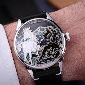 Op de afbeelding: Zilveren polshorloge met een zwarte leren band. De wijzerplaat toont een gedetailleerd ontwerp van twee paarden, één wit en één zwart, tegen een berglandschap. Het horloge maakt deel uit van de Travel Collection.