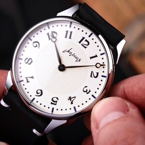 Könnte beinhalten: Nahaufnahme einer Armbanduhr mit weißem Zifferblatt, schwarzen Ziffern und schwarzen Zeigern. Die Uhr hat ein silberfarbenes Gehäuse und ein schwarzes Lederarmband. Auf dem Zifferblatt steht das Wort "Слава" in Schreibschrift.