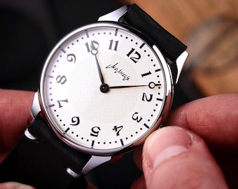 Könnte beinhalten: Nahaufnahme einer Armbanduhr mit weißem Zifferblatt, schwarzen Ziffern und schwarzen Zeigern. Die Uhr hat ein silberfarbenes Gehäuse und ein schwarzes Lederarmband. Auf dem Zifferblatt steht das Wort "Слава" in Schreibschrift.