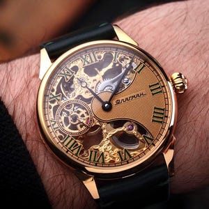 Op de afbeelding: Goudkleurig polshorloge met een skeleton wijzerplaat, Romeinse cijfers en ingewikkelde interne mechanismen. Het horloge heeft een zwarte leren band en een goudkleurige kast. De wijzerplaat toont het woord "Флагман".