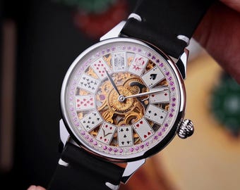 May include: A silver-toned wristwatch with a black leather band. The watch face features a playing card design with a gold-colored inner mechanism and pink accents. The watch is held by a person's hand.