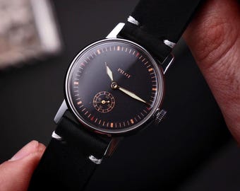 May include: A black wristwatch with a leather band is held by a person. The watch face is black with white hands and the word "PILOT" in white. The watch has a small seconds dial and a silver case.