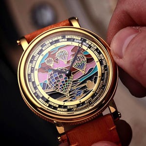 Op de afbeelding: Goudkleurig polshorloge met een bruine leren band. De wijzerplaat van het horloge heeft een kleurrijk luchtballonontwerp met stadsnamen rondom. Het horloge wordt door een hand vastgehouden.