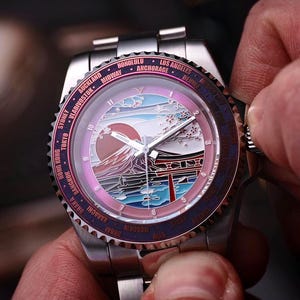 May include: A stainless steel wristwatch with a detailed dial featuring a Japanese landscape with Mount Fuji and a red sun. The watch has a rotating bezel with city names and is being adjusted by a person.