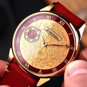 Könnte beinhalten: Eine goldfarbene Armbanduhr mit rotem Lederarmband. Das Zifferblatt der Uhr zeigt ein goldenes Blumenmuster und ein kleineres Zifferblatt. Römische Ziffern markieren die Stunden, und die Zeiger sind goldfarben. Die Uhr wird von einer Person gehalten.