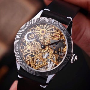 May include: A silver and gold skeleton watch with a black leather band. The watch face features intricate gears and a sunburst design. The watch is held by a person.