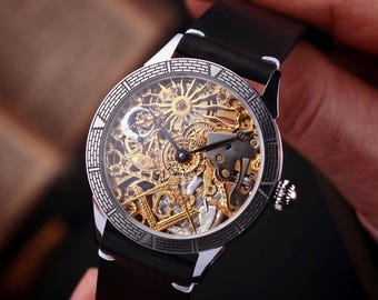 May include: A silver and gold skeleton watch with a black leather band. The watch face features intricate gears and a sunburst design. The watch is held by a person.