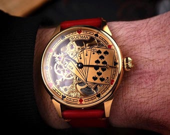 May include: Gold-toned wristwatch with a red leather band. The watch face features playing cards, including the ten of spades, and intricate mechanical details. The watch is on a wrist.