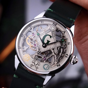 Op de afbeelding: Een zilverkleurig polshorloge met een groene leren band. De wijzerplaat heeft een skeletontwerp met vrijmetselaarsymbolen, waaronder een kompas, een vierkant en de letter "G". Het horloge heeft een gedetailleerd mechanisch uurwerk.