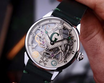 May include: A silver-toned wristwatch with a green leather strap. The watch face features a skeleton design with Masonic symbols, including a compass, square, and the letter "G". The watch has a detailed mechanical movement visible through the face.